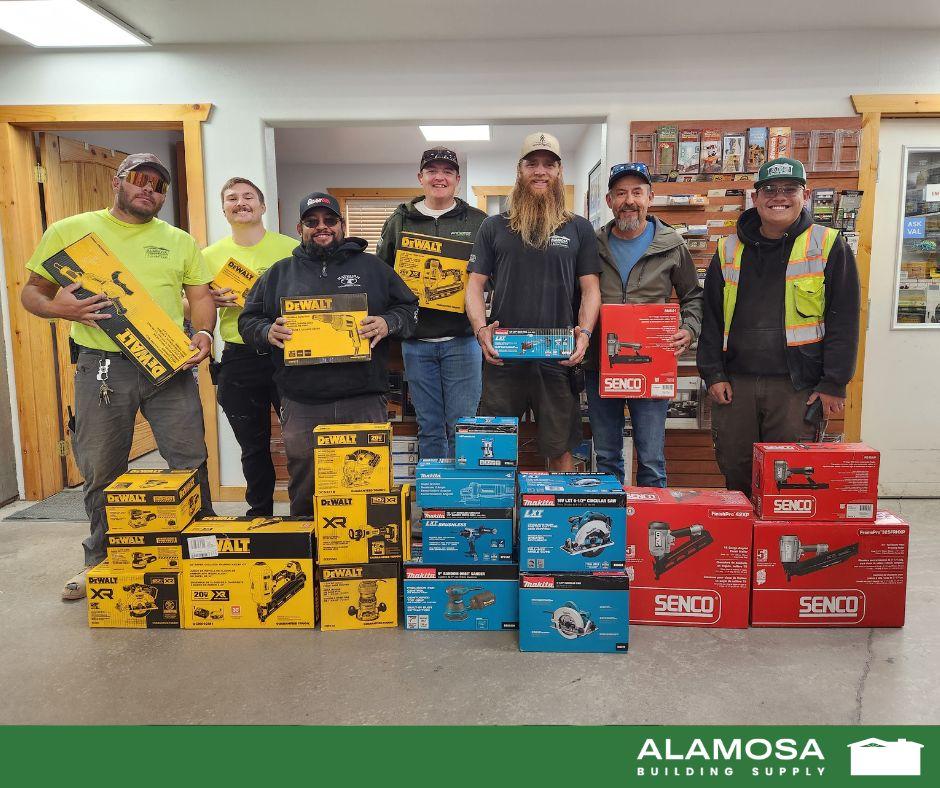 Construction crew members posing indoors with stacked DeWalt, Makita, and Senco tool boxes at Alamosa Building Supply.